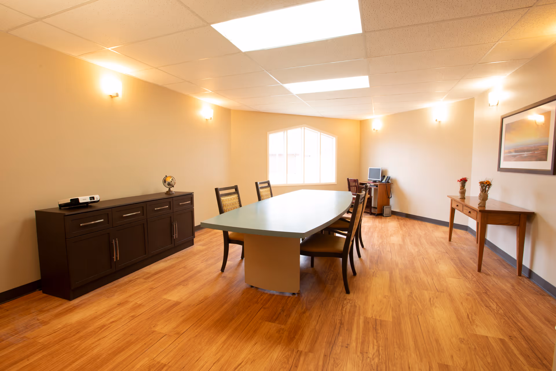 A well-lit conference or meeting room with a long oval table surrounded by six chairs. The room has wooden flooring, beige walls, and a window at the far end letting in natural light. There is a dark wooden cabinet on the left side with a small globe and a projector on top. On the right side, there is a wooden console table with two small vases of flowers and a framed picture hanging above it. A desk with a computer and chair is positioned near the window.