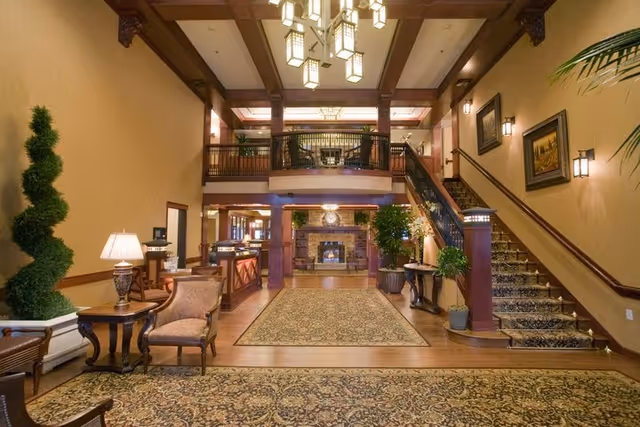 Spacious and elegant interior of a senior living facility with a large patterned carpet, wooden furniture including chairs and tables, potted plants, a staircase with a carpet runner, and a fireplace in the background under a high ceiling with hanging lights.