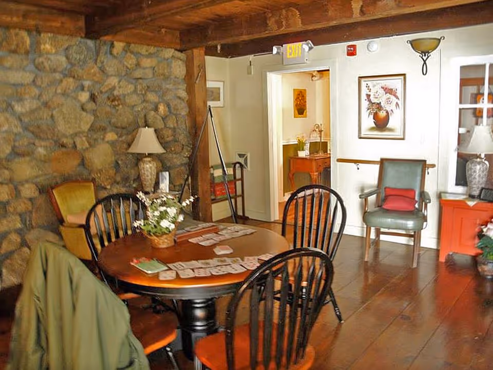 A cozy interior room with a round wooden table surrounded by black wooden chairs. The table has playing cards and a small flower arrangement on it. The room features a stone wall, wooden floor, and ceiling beams. There are two lamps on side tables, a green armchair with a red cushion, framed artwork on the walls, and an open doorway leading to another room.