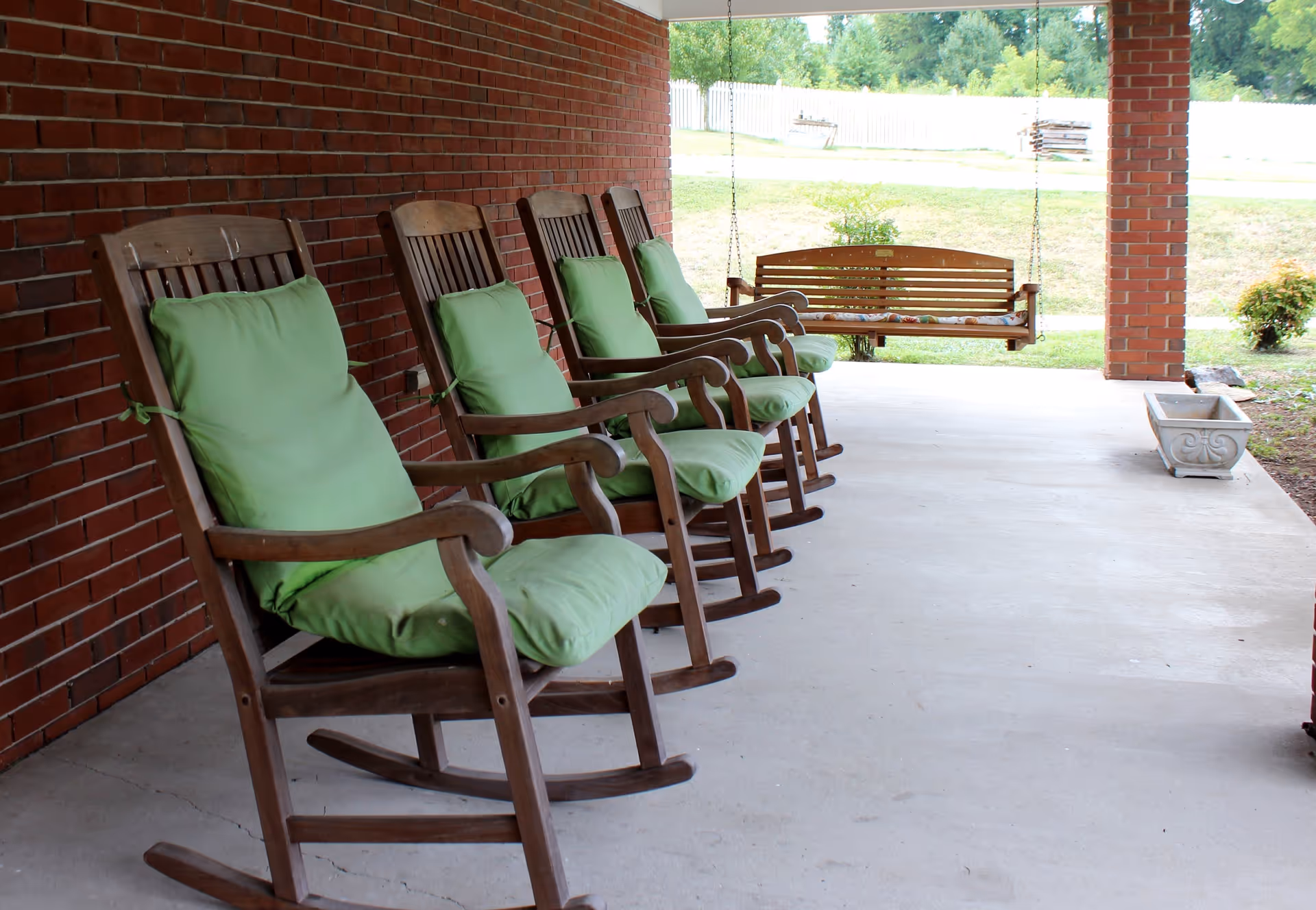 A covered outdoor patio area with five wooden rocking chairs lined up against a brick wall, each with green cushions. At the end of the patio, there is a wooden porch swing hanging from the ceiling. The patio overlooks a grassy area with some bushes and a white fence in the background.