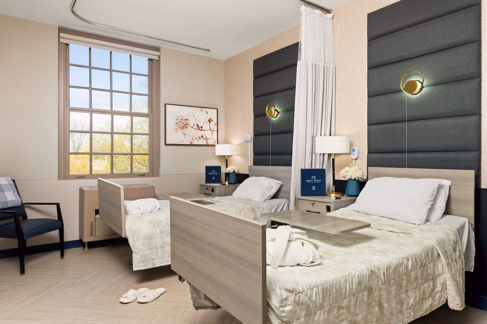 A bright and clean shared bedroom in a healthcare facility featuring two single beds with light wood frames and padded dark gray headboards. Each bed has white pillows and cream-colored bedding. Between the beds is a small nightstand with a lamp, flowers, and a Liberty Pointe Rehabilitation and Healthcare Center booklet. A privacy curtain hangs between the beds. There is a large window with a view of trees, a chair with a plaid blanket, and slippers on the light wood floor.