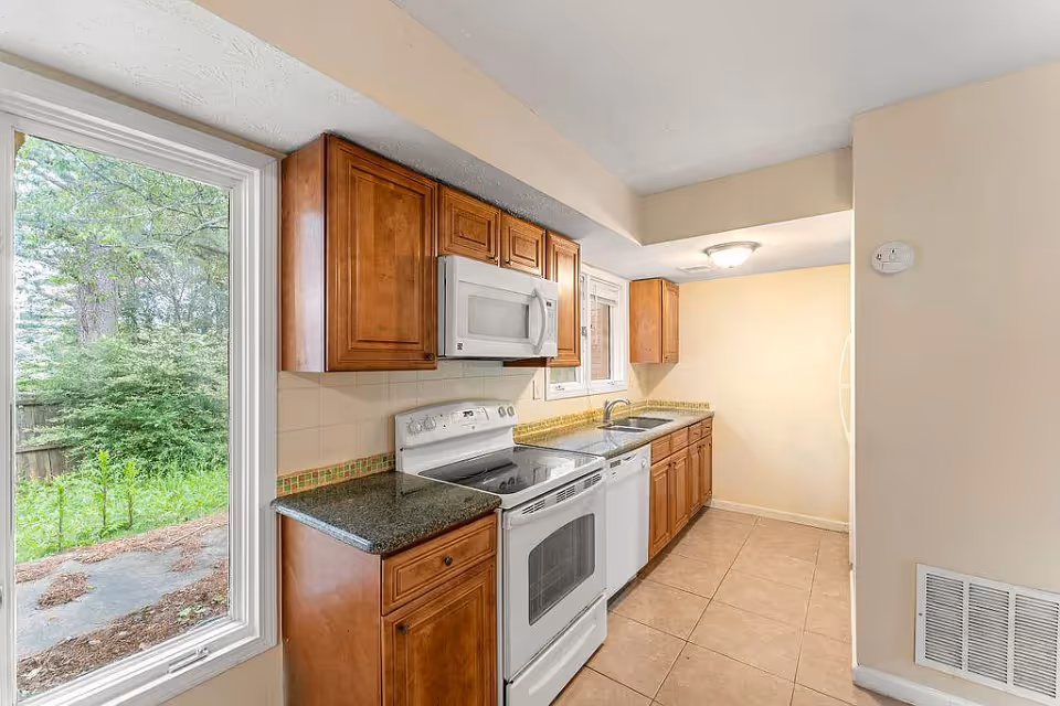A bright kitchen with wooden cabinets, a white electric stove with oven, a white microwave above the stove, a dishwasher, and a double sink. There is a large window on the left side showing green trees and bushes outside. The floor is tiled and the walls are painted beige.