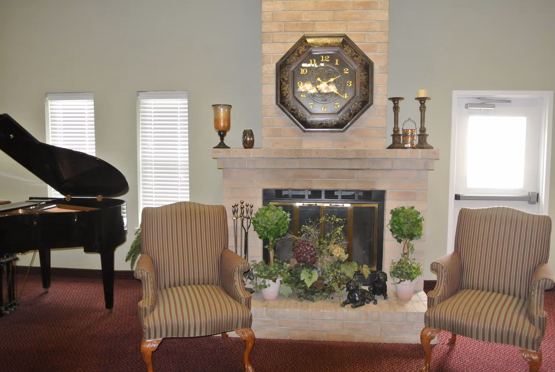 Two striped upholstered armchairs flank a brick fireplace topped with a decorative clock and plants, with a grand piano visible to the left.