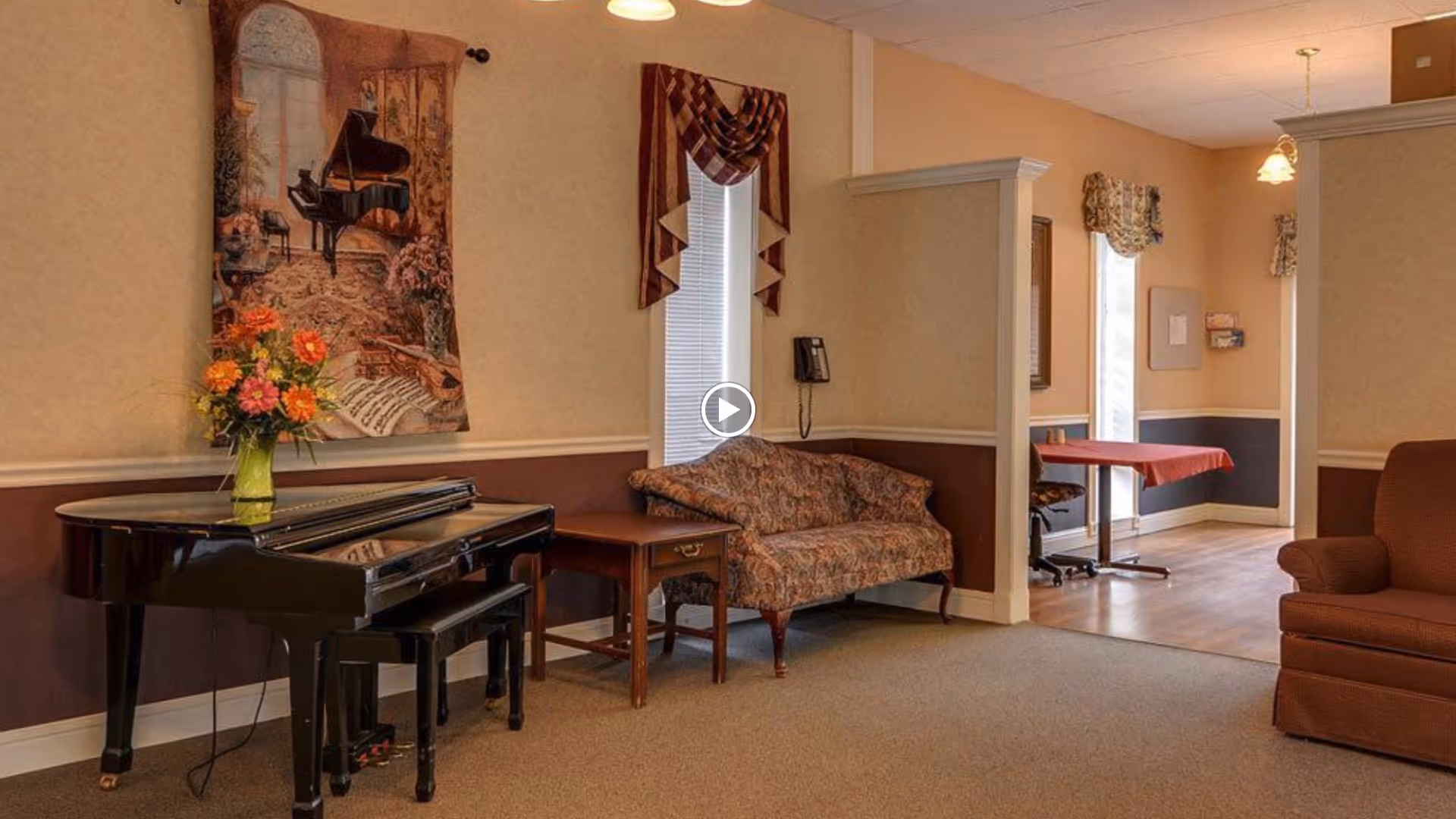 Interior view of a nursing center common area featuring a black grand piano with a vase of flowers on top, a floral-patterned loveseat, a wooden side table, and a brown armchair. The walls are beige with a darker brown wainscoting, decorated with a tapestry of a piano scene and window treatments with valances. In the background, there is a small table with a red tablecloth and a chair near a window.
