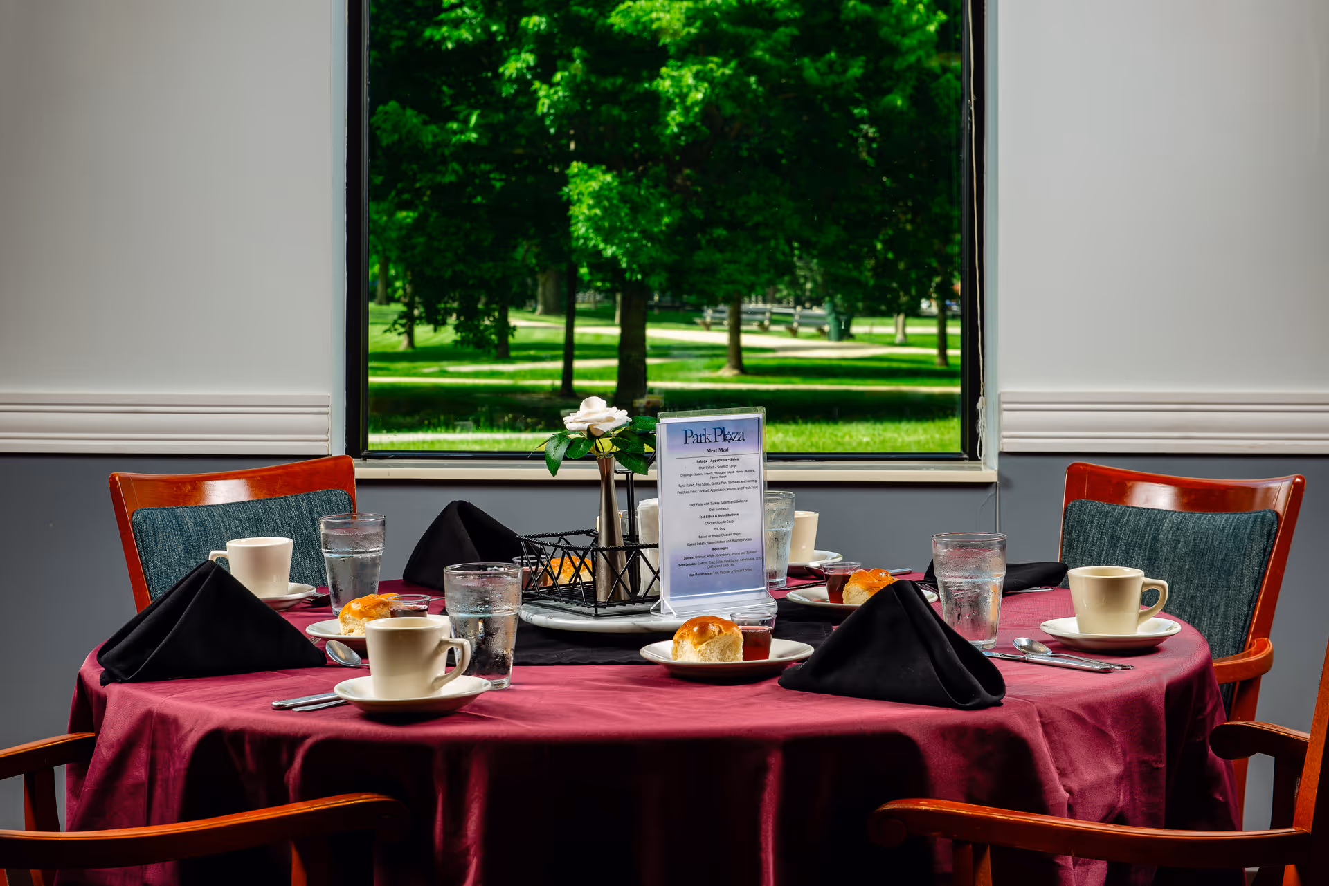 A round dining table covered with a burgundy tablecloth set for four people with black folded napkins, cups, glasses of water, bread rolls, and a small menu card. The table is positioned in front of a window showing a green outdoor park with trees and a walking path.