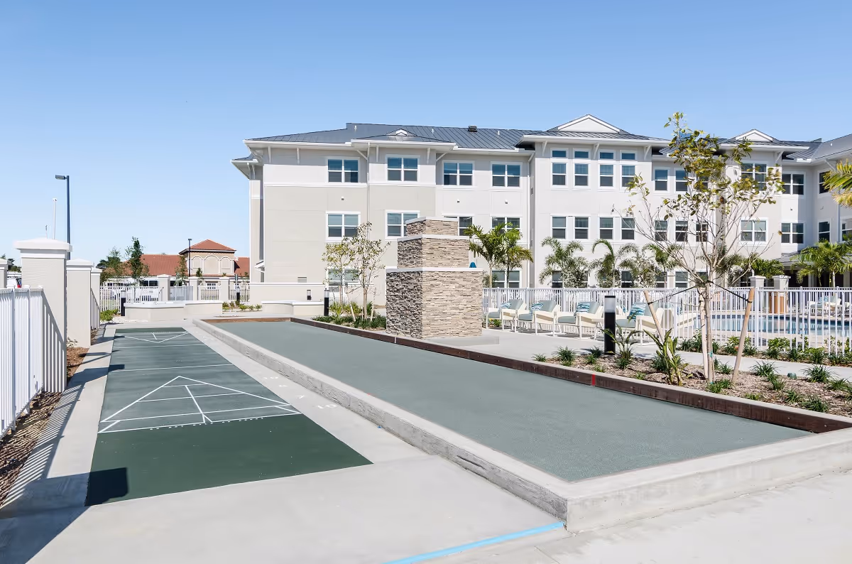 Outdoor recreational area at American House Ft. Myers featuring a shuffleboard court and bocce ball court with seating and landscaping, adjacent to a multi-story residential building under clear blue sky.