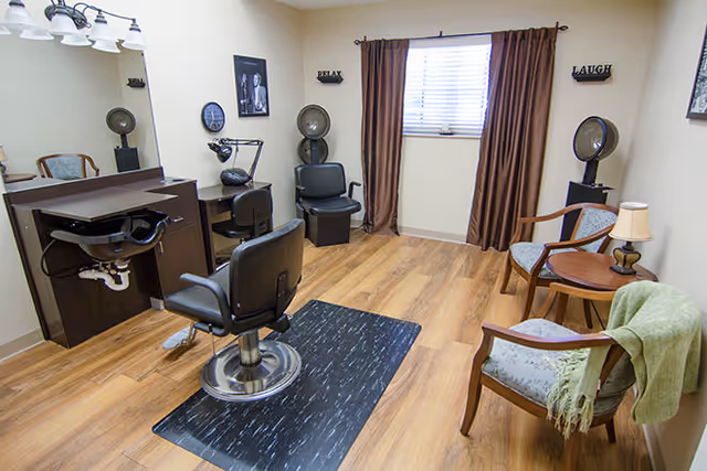 A small hair salon room with a styling chair in front of a mirror and a hair washing station. The room has wooden flooring, a window with brown curtains, two chairs with cushions, a small round table with a lamp, and two vintage hair dryers. Wall decorations include framed pictures and signs that say 'RELAX' and 'LAUGH'.