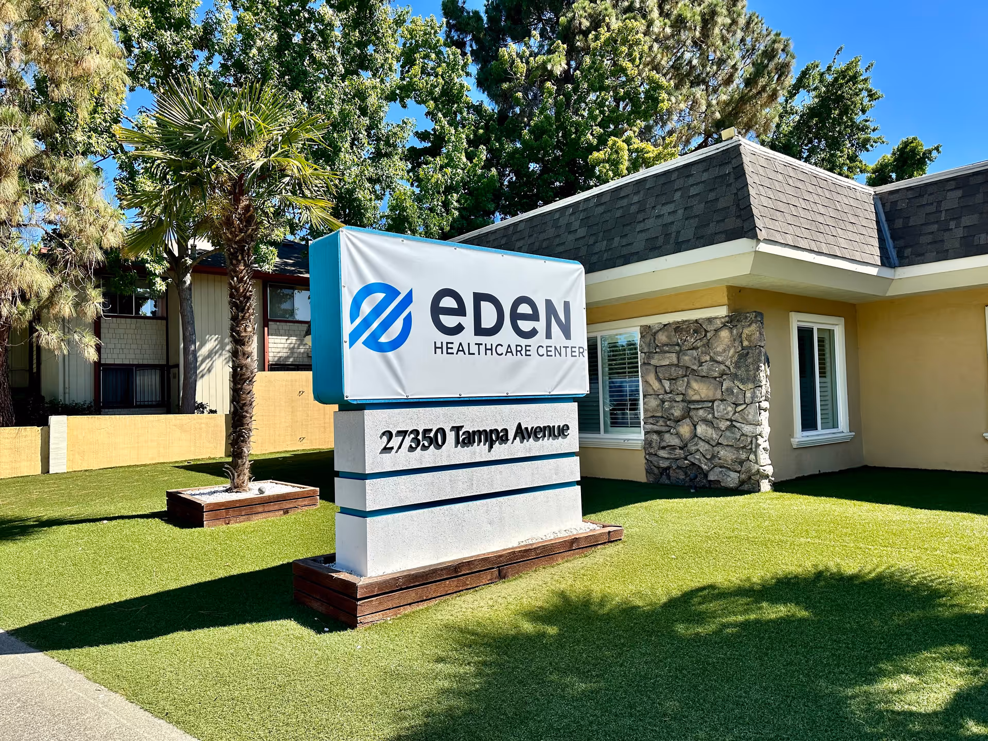 Outdoor view of Eden Healthcare Center with a sign displaying the facility's name and address, 27350 Tampa Avenue, surrounded by green grass, palm trees, and other trees under a clear blue sky.