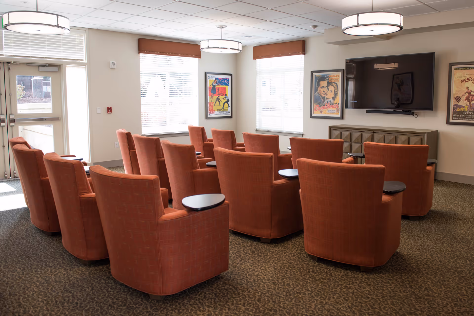 A small media room with rows of orange upholstered armchairs facing a wall-mounted TV and framed posters.
