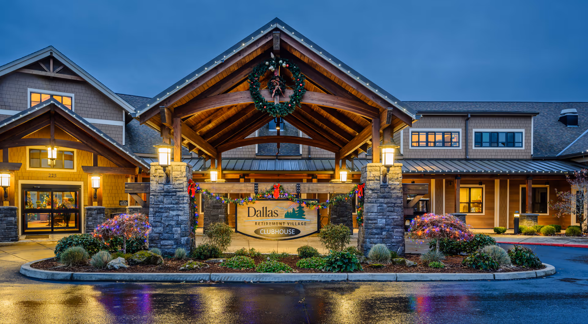 Exterior view of the Dallas Retirement Village clubhouse entrance with a wooden portico, stone pillars, festive wreaths, and landscaped shrubs at dusk.