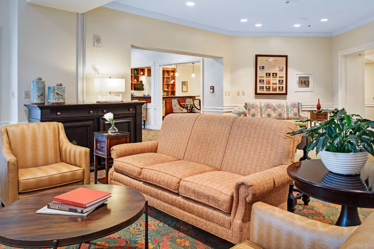 A cozy living room area in a senior living facility featuring a brown upholstered sofa, two tan leather armchairs, a round wooden coffee table with books, a dark wooden sideboard with decorative vases and a lamp, and a round side table with a potted plant. The room has light-colored walls, patterned carpet, and framed pictures on the walls. An open doorway leads to another room with wooden shelves and additional seating.