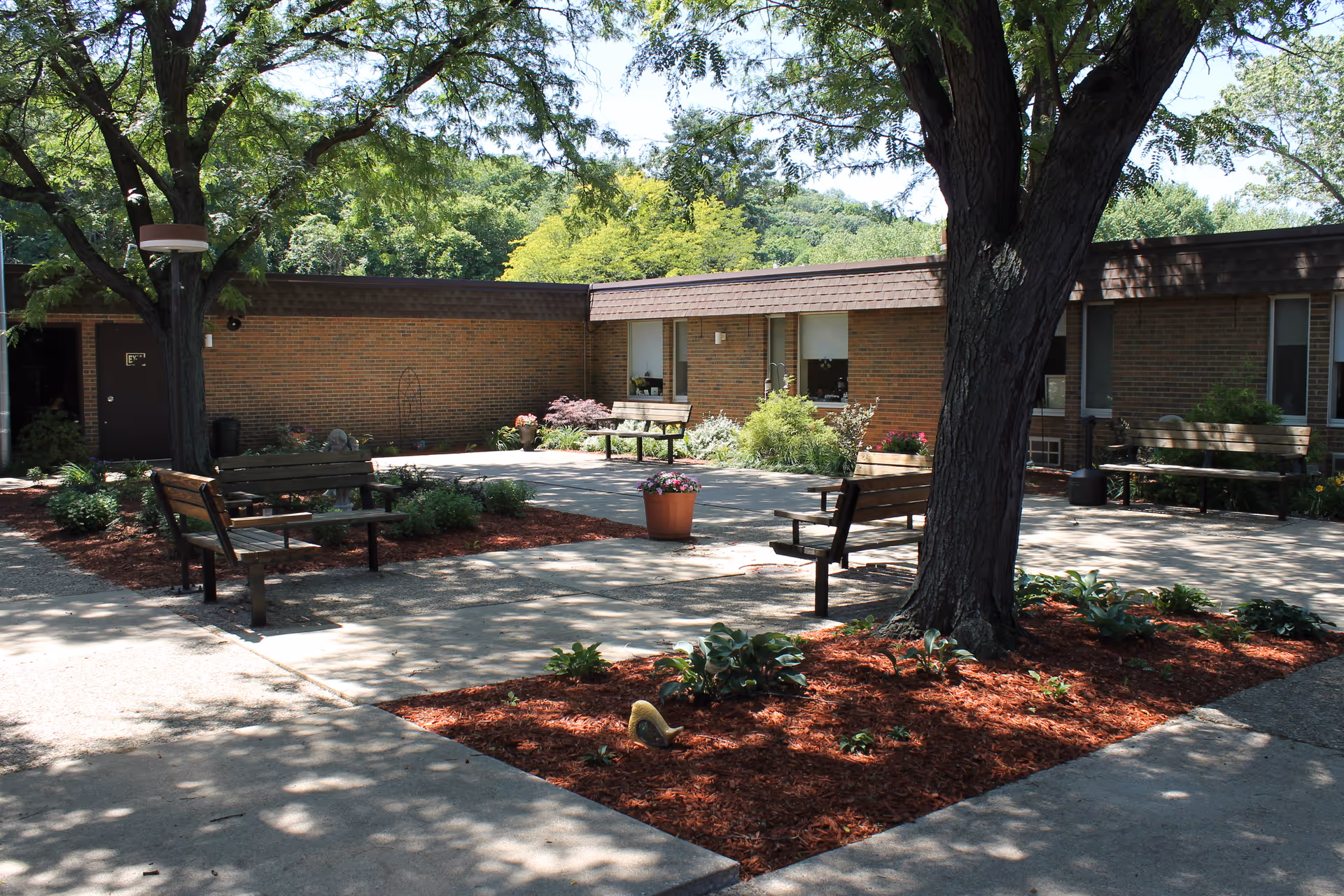 Outdoor courtyard area at Goldwater Care Peoria Heights featuring several wooden benches, a large tree providing shade, landscaped garden beds with mulch and plants, and a brick building in the background under a clear sky.
