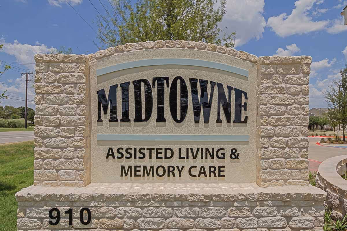 Stone monument sign for Midtowne Assisted Living & Memory Care with the number 910 at the base, set outdoors with a blue sky and some trees in the background.