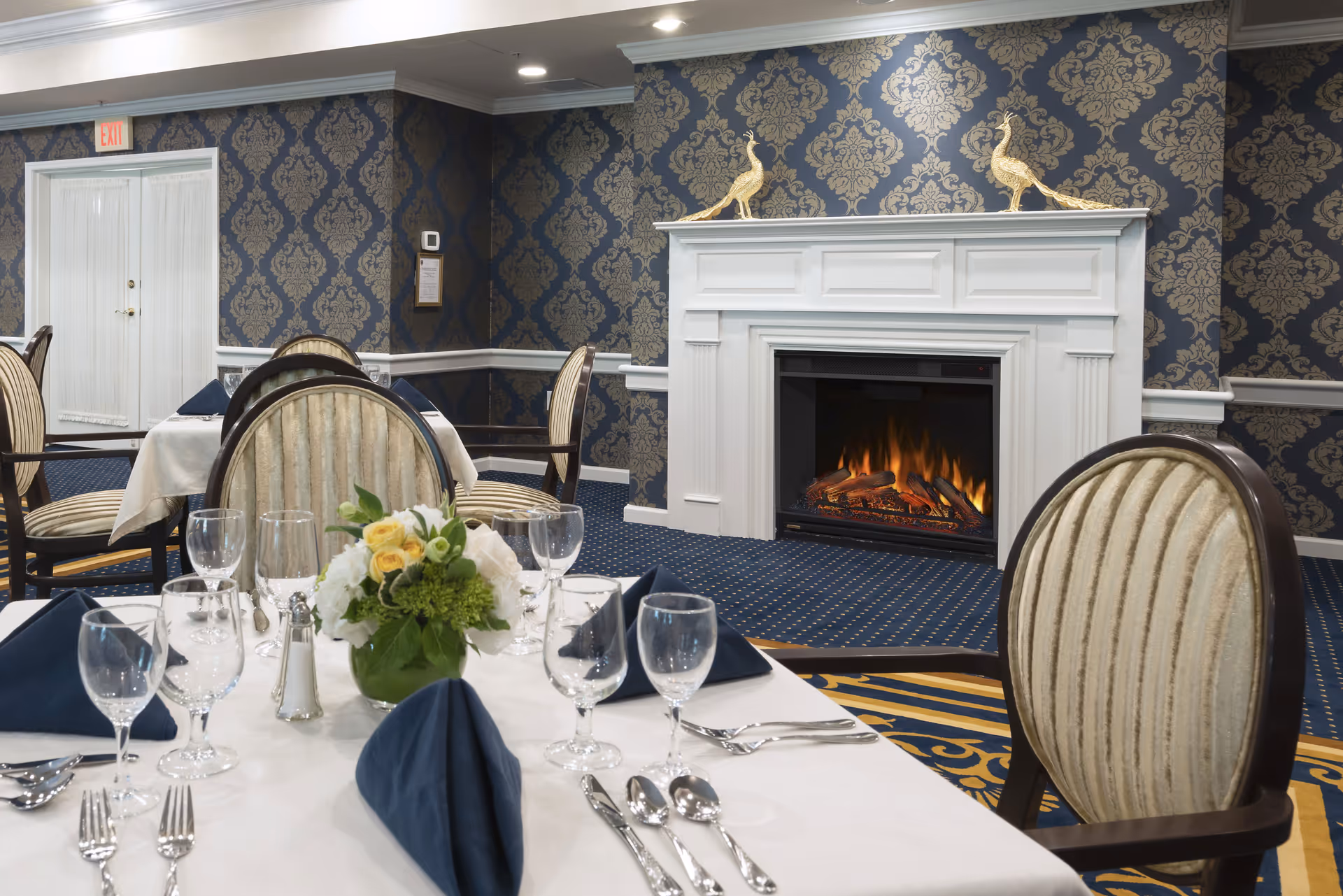 Elegant dining room with tables set with white tablecloths, blue napkins, glassware, and silverware. A floral centerpiece is on the table in the foreground. The room features a white fireplace with a fire burning and two golden peacock statues on the mantel. The walls have blue and gold damask wallpaper, and the carpet is blue with a patterned design.