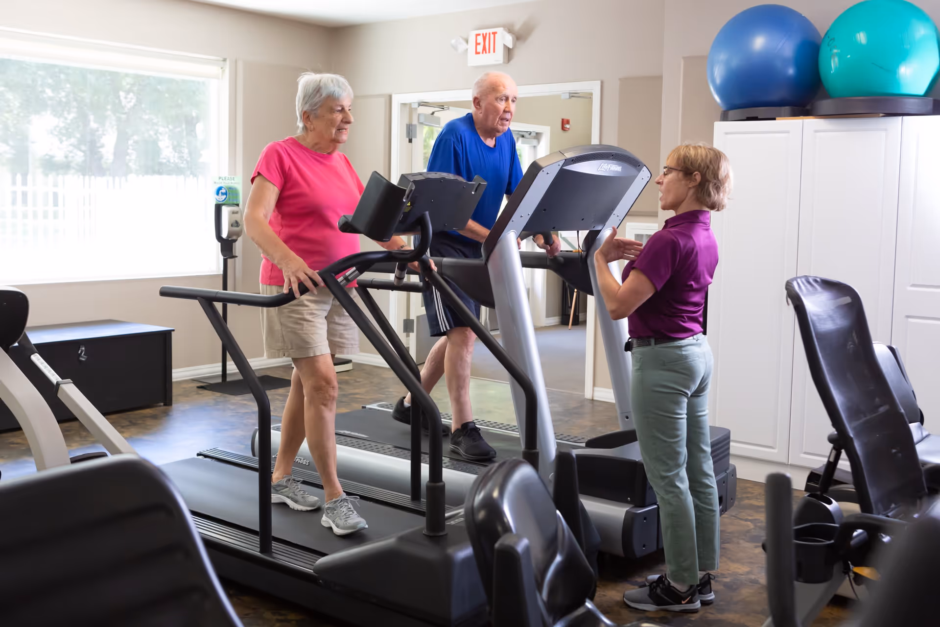 Two elderly individuals exercising on treadmills in a fitness room while a fitness instructor stands nearby giving guidance. The room has exercise equipment, large windows, and exercise balls on top of a cabinet.