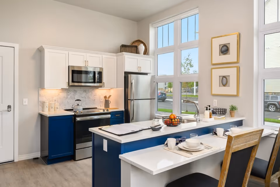 Modern kitchen with white upper cabinets and blue lower cabinets, stainless steel appliances including a microwave, stove, and refrigerator. A kitchen island with a sink and a breakfast bar with two place settings is in the foreground. Large windows provide natural light and a view of the outside.