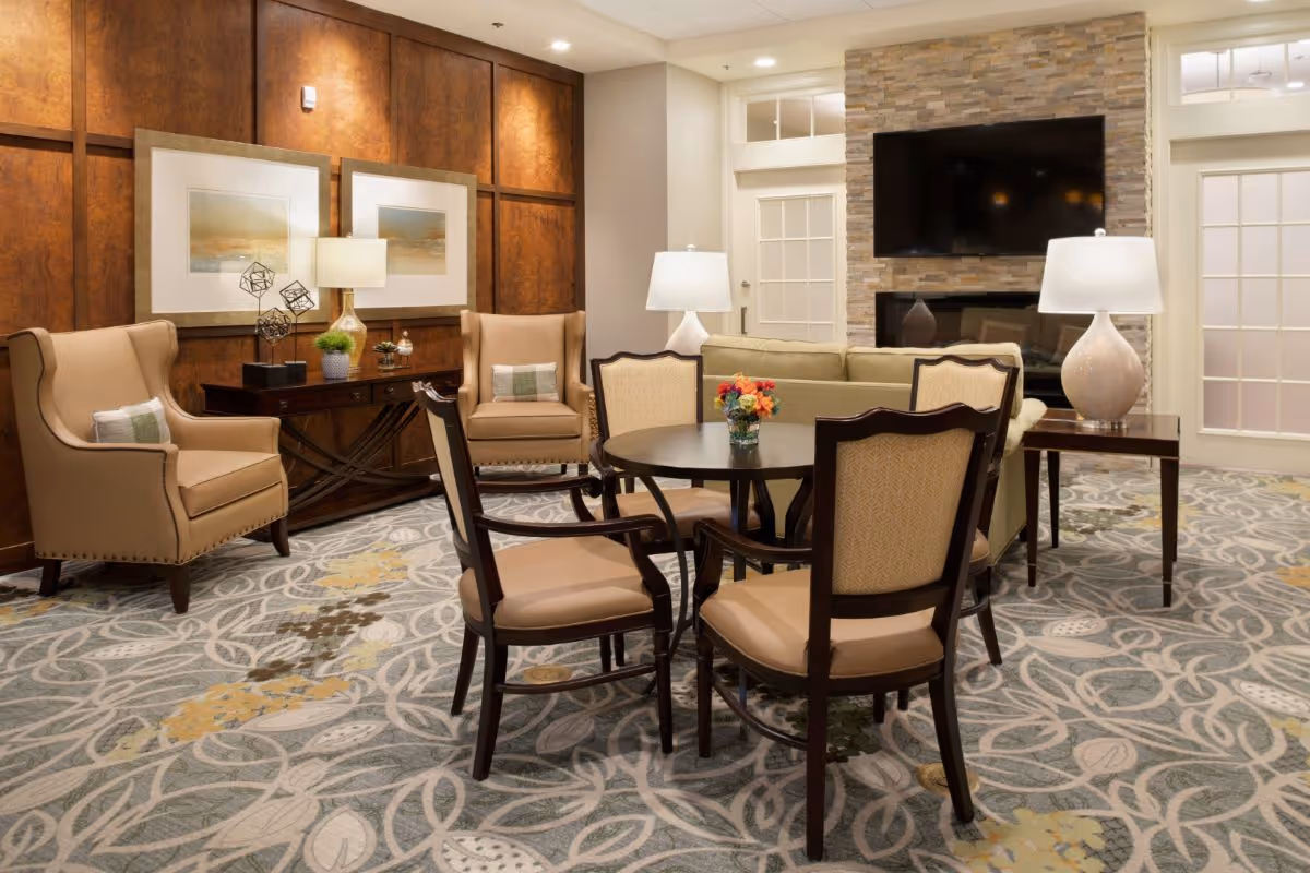 A cozy living room area with a round table surrounded by four upholstered chairs, two beige armchairs with pillows, a sofa, two table lamps on side tables, a wooden console table with decorative items, framed artwork on the wall, a patterned carpet, and a stone fireplace with a mounted flat-screen TV.