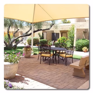 Outdoor patio area with a round metal table and four chairs with yellow cushions under a large beige canopy. The patio is paved with bricks and surrounded by potted plants, bushes, and trees. Beige building walls and windows are visible in the background.