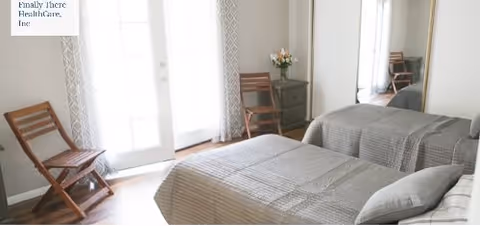 A bedroom with a bed covered in a gray quilt, a wooden folding chair, a small green nightstand with a vase of flowers, and a large mirror reflecting part of the room. There are glass doors with patterned curtains allowing natural light to enter.