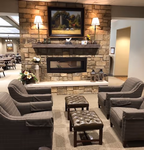 A cozy seating area in a senior living facility with four gray upholstered armchairs arranged around two tufted leather ottomans. The background features a stone fireplace with a wooden mantel, decorated with two table lamps, small decorative items, and a framed painting of a rural scene with sheep. To the left, there is a floral arrangement and a glimpse of a dining area with tables and chairs.