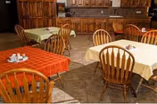 Dining area with several tables covered in colorful tablecloths and wooden chairs in front of a wall of kitchen cabinets.