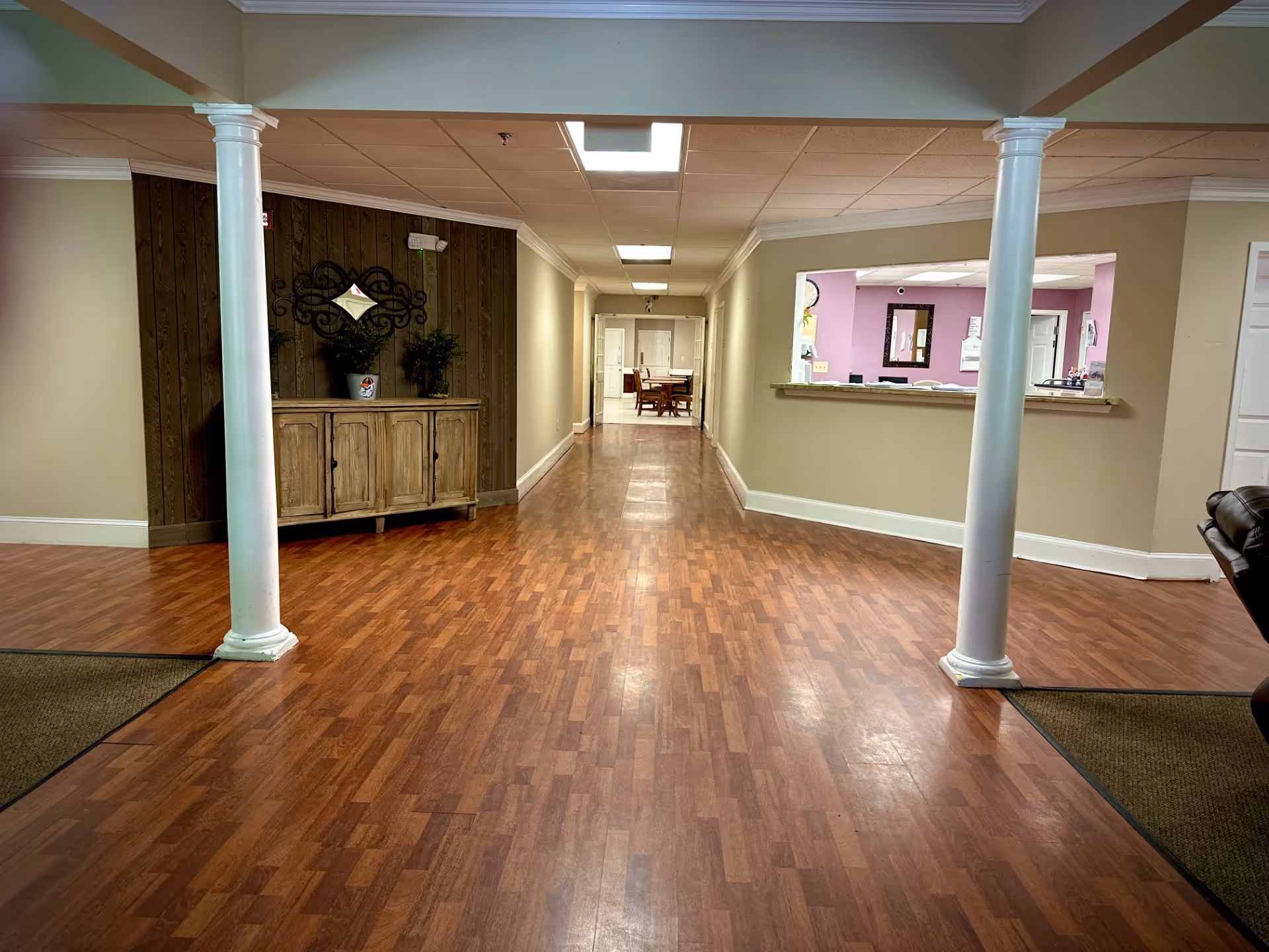 Interior hallway of a senior living facility with wooden flooring, two white columns, a wooden cabinet with plants and a decorative mirror on the left wall, and a reception window with a purple wall behind it on the right. At the end of the hallway, there is a table with chairs.