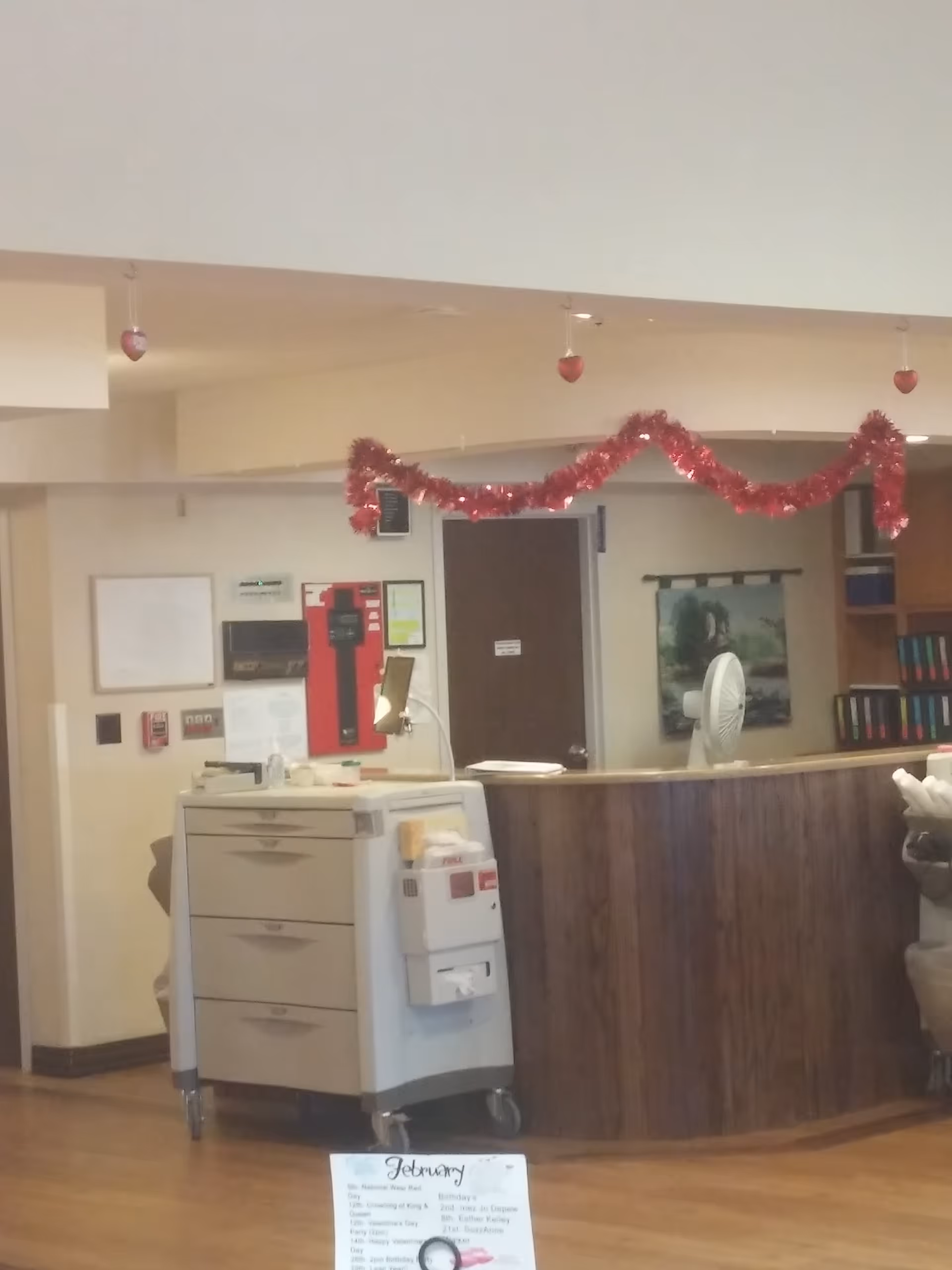 Reception area inside a senior living facility with a curved wood desk, a rolling cart, and red garland decorations.