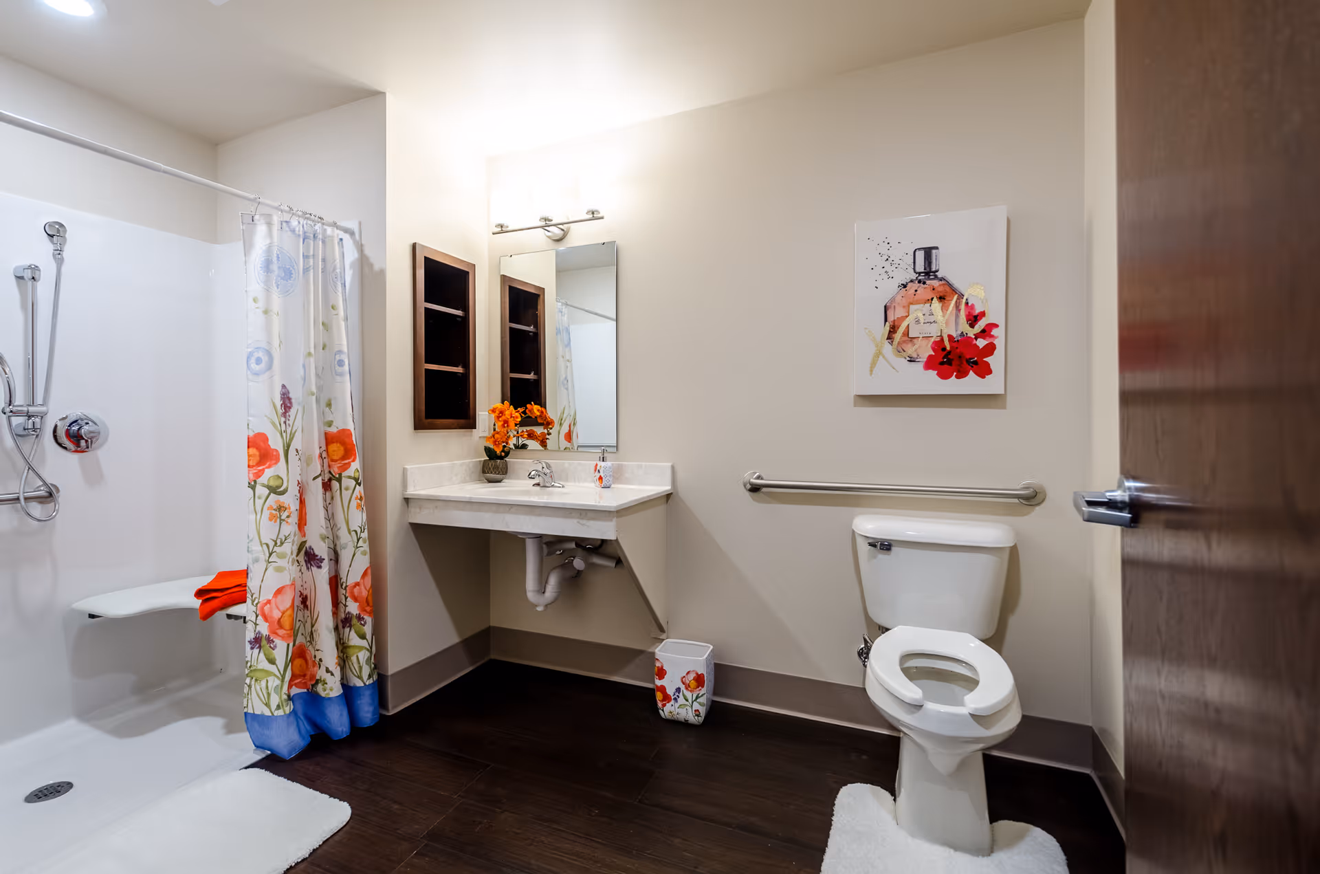 A clean and accessible bathroom with a white toilet, a wall-mounted sink with a mirror above it, a floral shower curtain, and a small trash bin with floral design. There is a grab bar next to the toilet and a piece of wall art featuring a perfume bottle and red flowers. The floor is dark wood, and the walls are light beige.