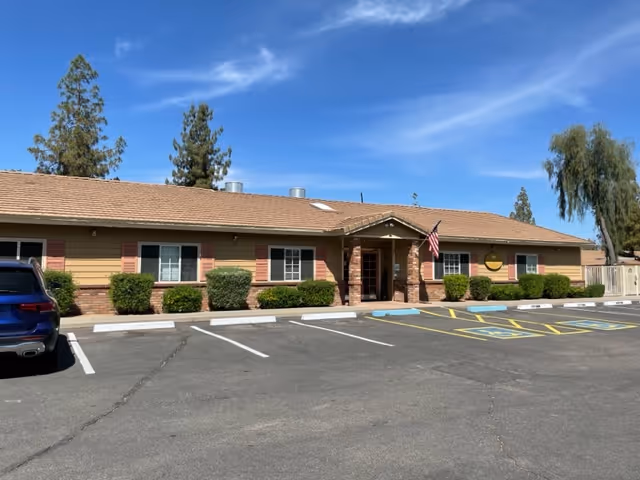 Single-story building with a brown roof and beige siding with red shutters, surrounded by bushes and a parking lot with marked spaces including handicapped spots. An American flag is displayed near the entrance under a small covered porch. The sky is clear and blue with a few wispy clouds.