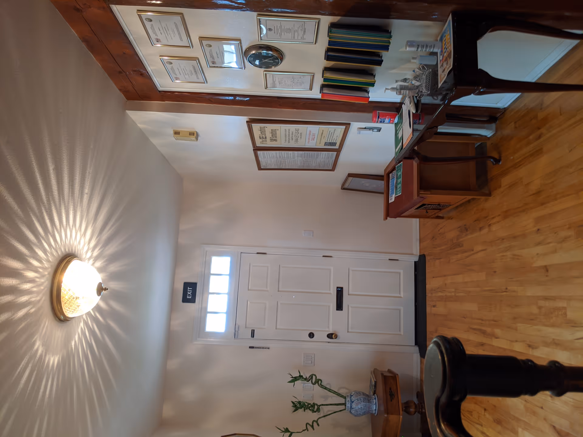 Interior hallway of a senior living facility with a wooden floor, a white exit door with a small window above it, a ceiling light fixture casting decorative shadows, a small wooden table with a blue and white vase holding bamboo stalks, a wooden bench, and a wall with framed certificates, a clock, and informational pamphlets.