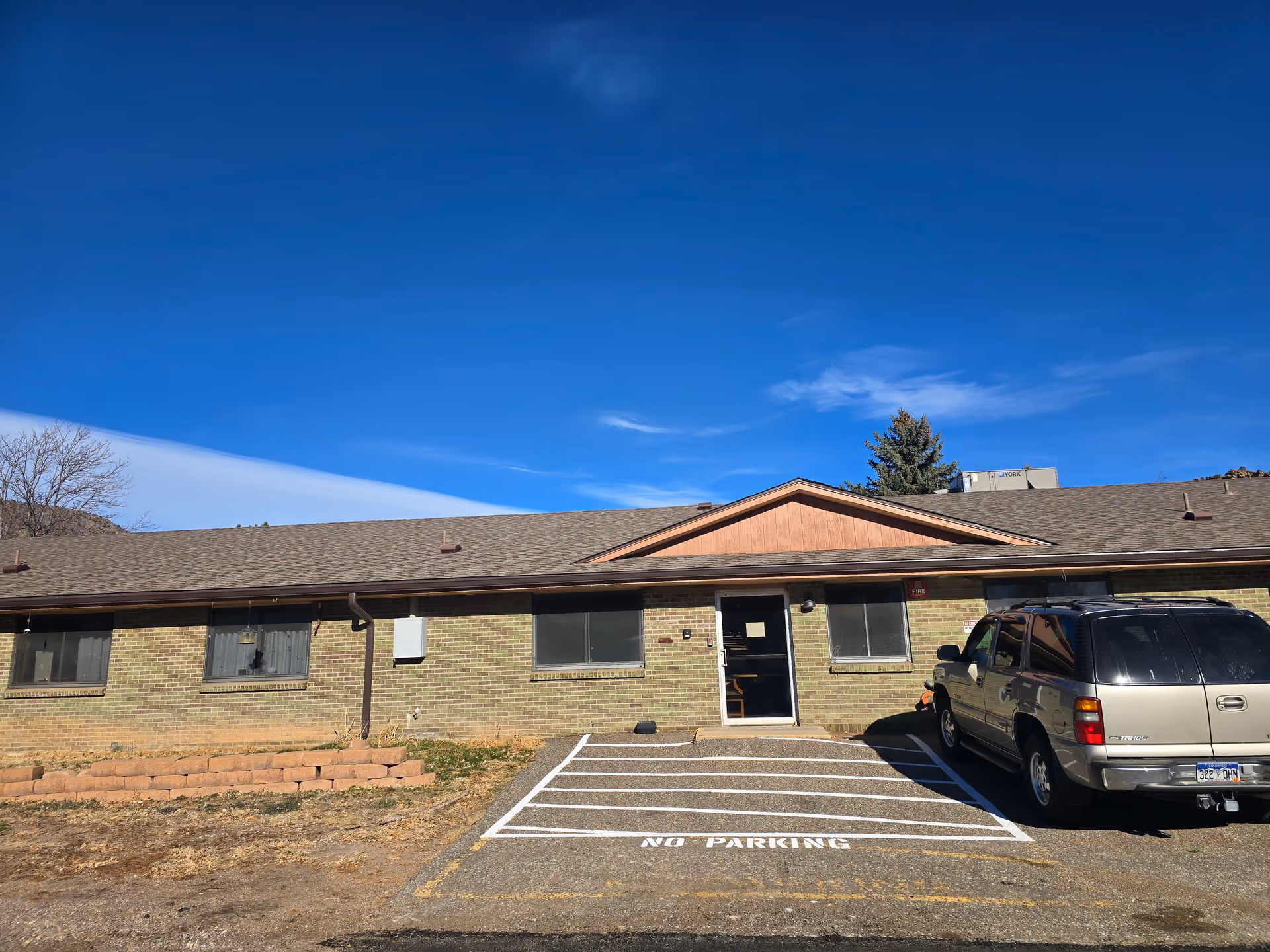 Exterior view of a single-story brick building under a clear blue sky with a parked silver SUV in front. The building has several windows, a central entrance door, and a 'NO PARKING' zone marked on the pavement.