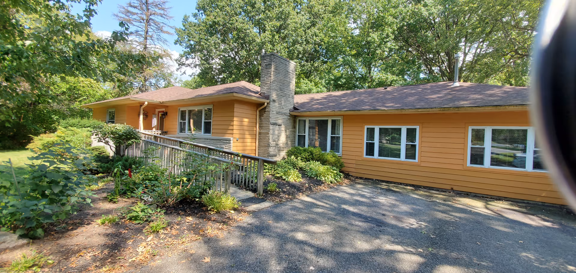 Exterior view of a single-story building with orange siding and a brown roof, surrounded by greenery and trees. There is a paved driveway in front and a wooden ramp leading to the entrance.