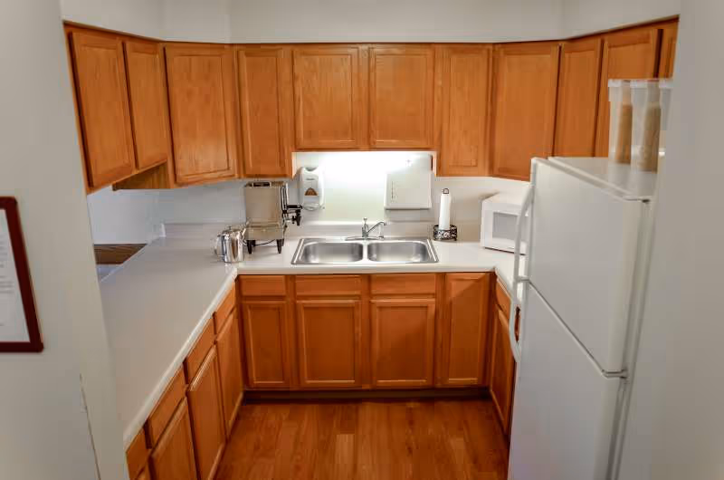 Small U-shaped kitchen with wooden cabinets, a double sink, under-cabinet lighting, countertop appliances, and a white refrigerator.