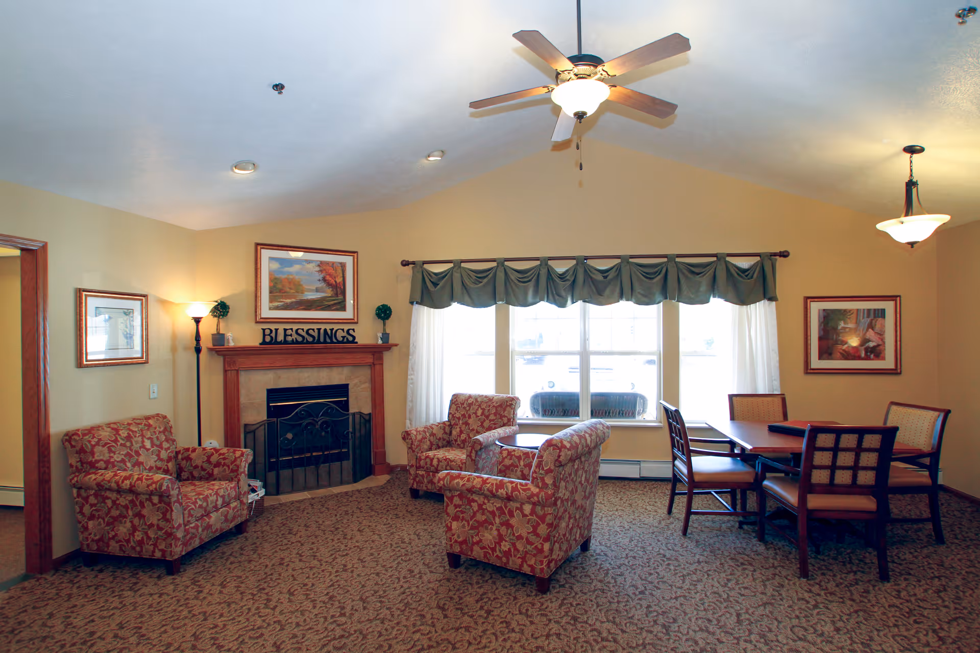 Sunlit common room with a fireplace, floral armchairs, a ceiling fan, and a dining table by a large window.