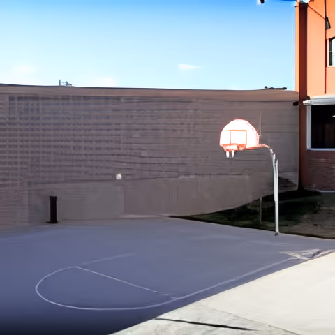 A small outdoor half-basketball court with a single hoop next to a tall brick and concrete building under a clear blue sky.