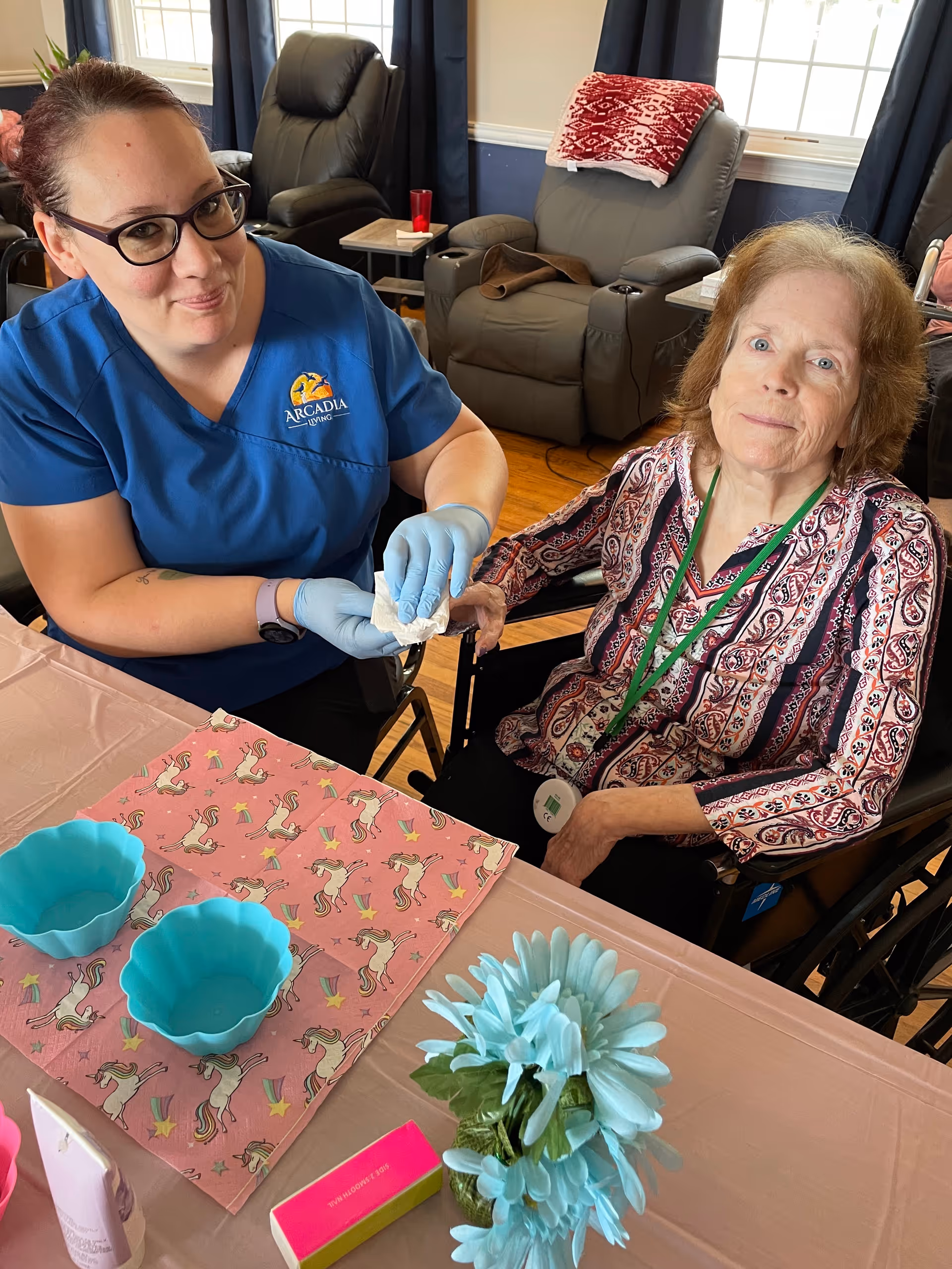 A caregiver wearing blue scrubs with the Arcadia Living logo and blue gloves is gently holding the hand of an elderly woman seated in a wheelchair. They are at a table covered with a pink tablecloth and a unicorn-patterned cloth, with blue silicone bowls, a flower arrangement, and nail care items visible. The setting appears to be a cozy living room with recliner chairs and windows in the background.