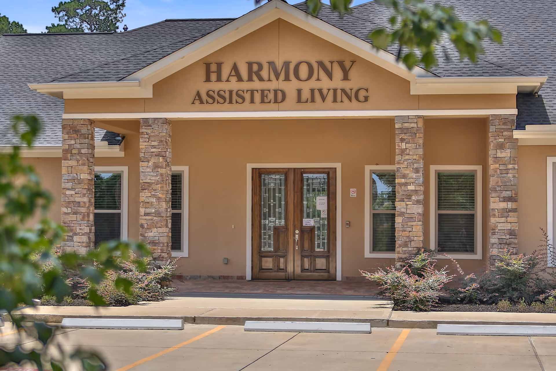Front exterior view of Harmony Assisted Living facility showing a beige building with stone pillars, a double wooden door entrance, and the facility name displayed above the entrance.