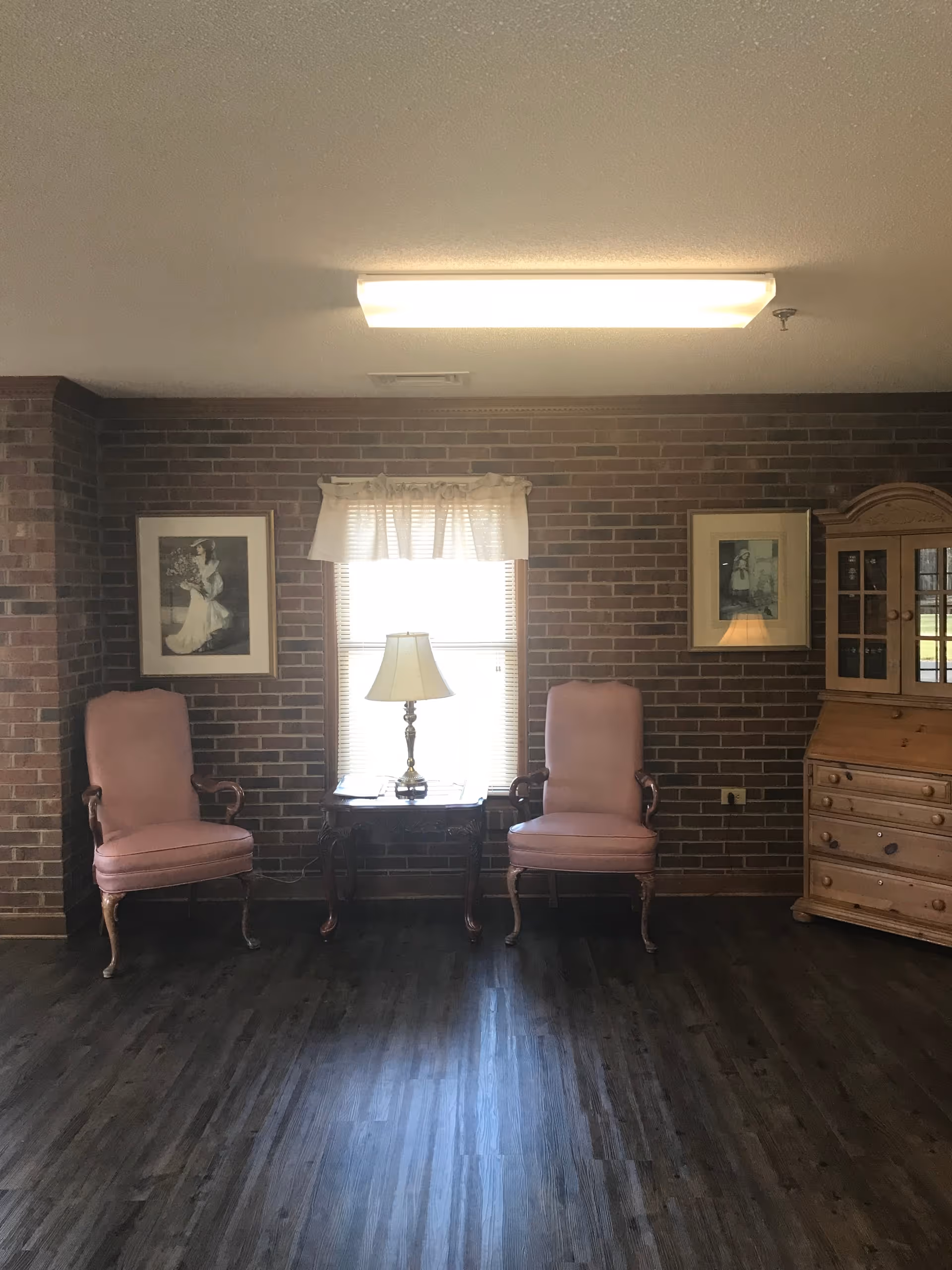 A sitting area with two pink upholstered armchairs placed on either side of a wooden side table with a lamp. The background features a brick wall with a window in the center, covered by a white valance. There are two framed black and white pictures hanging on the wall, one on each side of the window. To the right, there is a wooden cabinet with glass doors and drawers. The floor is dark wood.