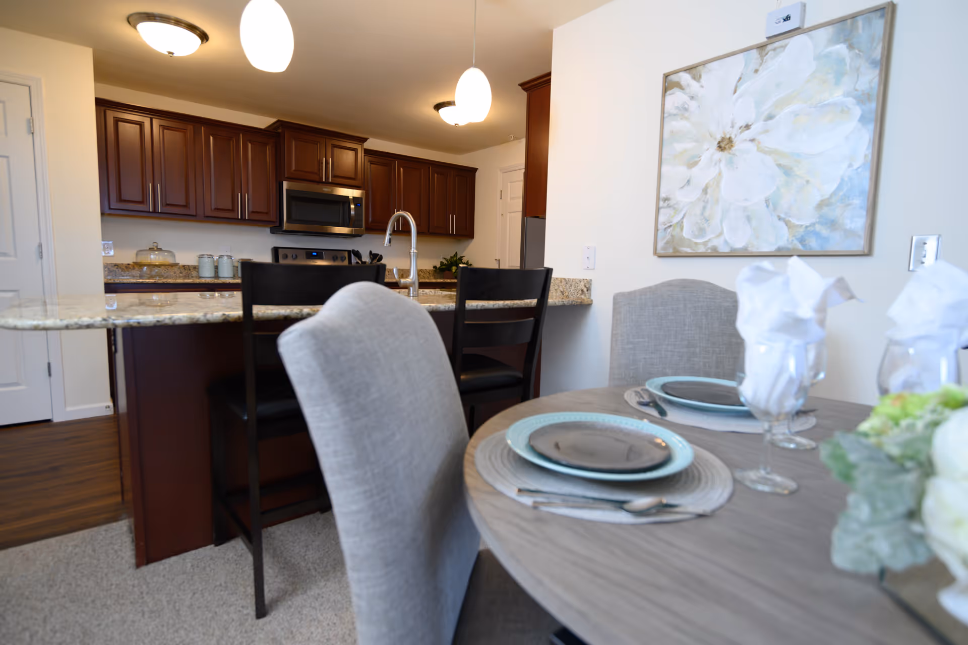 A modern kitchen and dining area in a senior living facility. The kitchen features dark wood cabinets, a stainless steel microwave and stove, and a granite countertop with a sink and two black bar stools. The dining area has a round wooden table set with plates, silverware, and glasses with white napkins, along with upholstered chairs. A large floral painting hangs on the wall above the dining table.