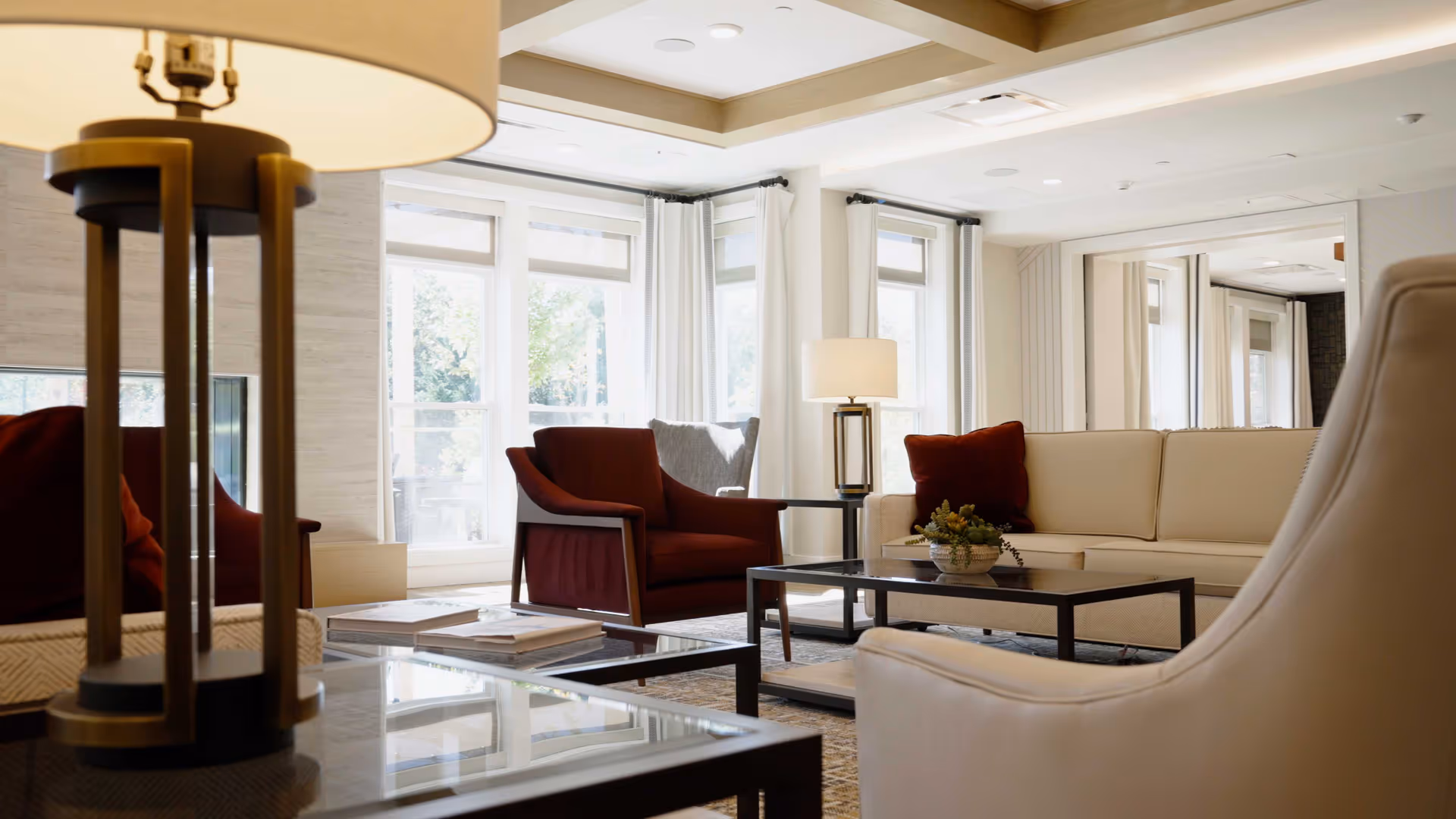 Well-lit senior living lounge with sofas, armchairs, coffee tables and lamps by large windows.