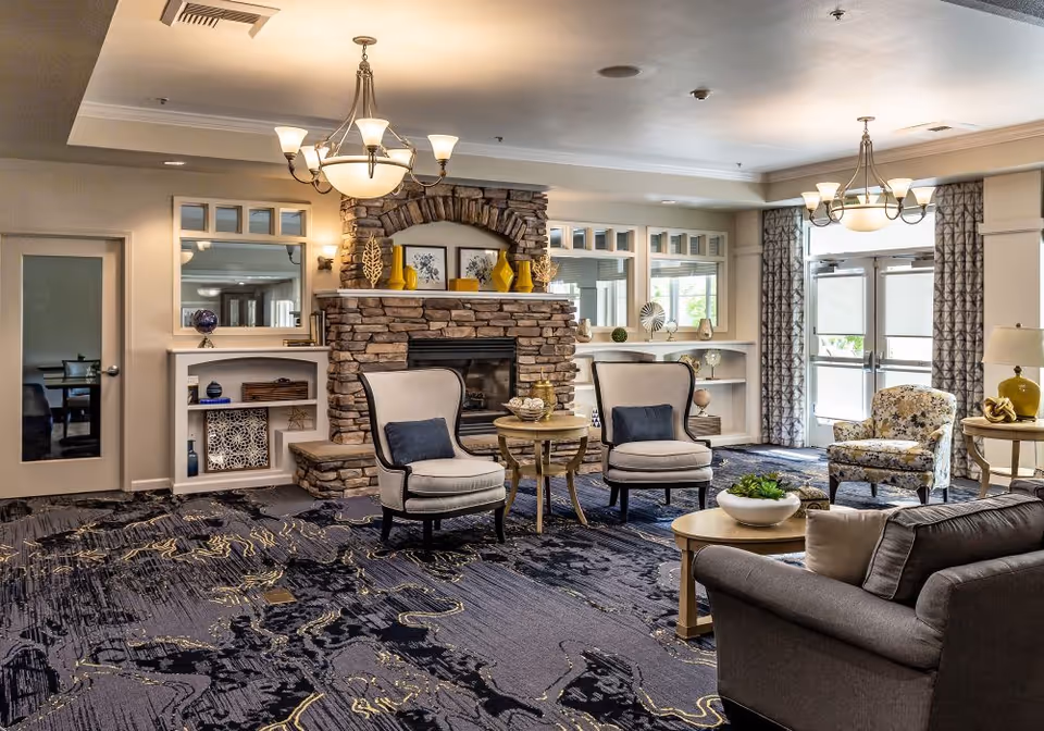 A cozy senior living facility common area with a stone fireplace as the focal point. The room features two beige armchairs with dark cushions, a round wooden table between them, a floral patterned armchair, and a gray sofa with pillows. The walls have built-in shelves decorated with vases and art pieces. Large windows and glass doors with patterned curtains allow natural light to enter. Two elegant chandeliers hang from the ceiling, and the carpet has a modern abstract design in shades of blue and gray.