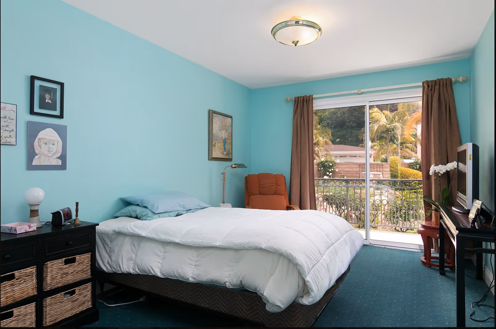 A bedroom with turquoise walls and a large window with brown curtains overlooking a garden with palm trees. The room has a bed with white bedding and light blue pillows, a black dresser with wicker baskets, a small lamp, and an alarm clock. There is an orange armchair in the corner and a TV on a black stand with a white orchid plant beside it. Several framed pictures and artwork hang on the walls.
