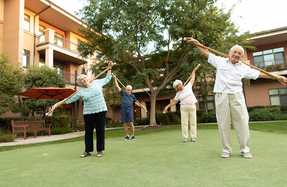 Four elderly people standing on a green lawn outside a retirement community building, each holding a wooden stick and stretching their arms to the side as part of a group exercise. The background shows trees, benches, and a multi-story building with balconies.