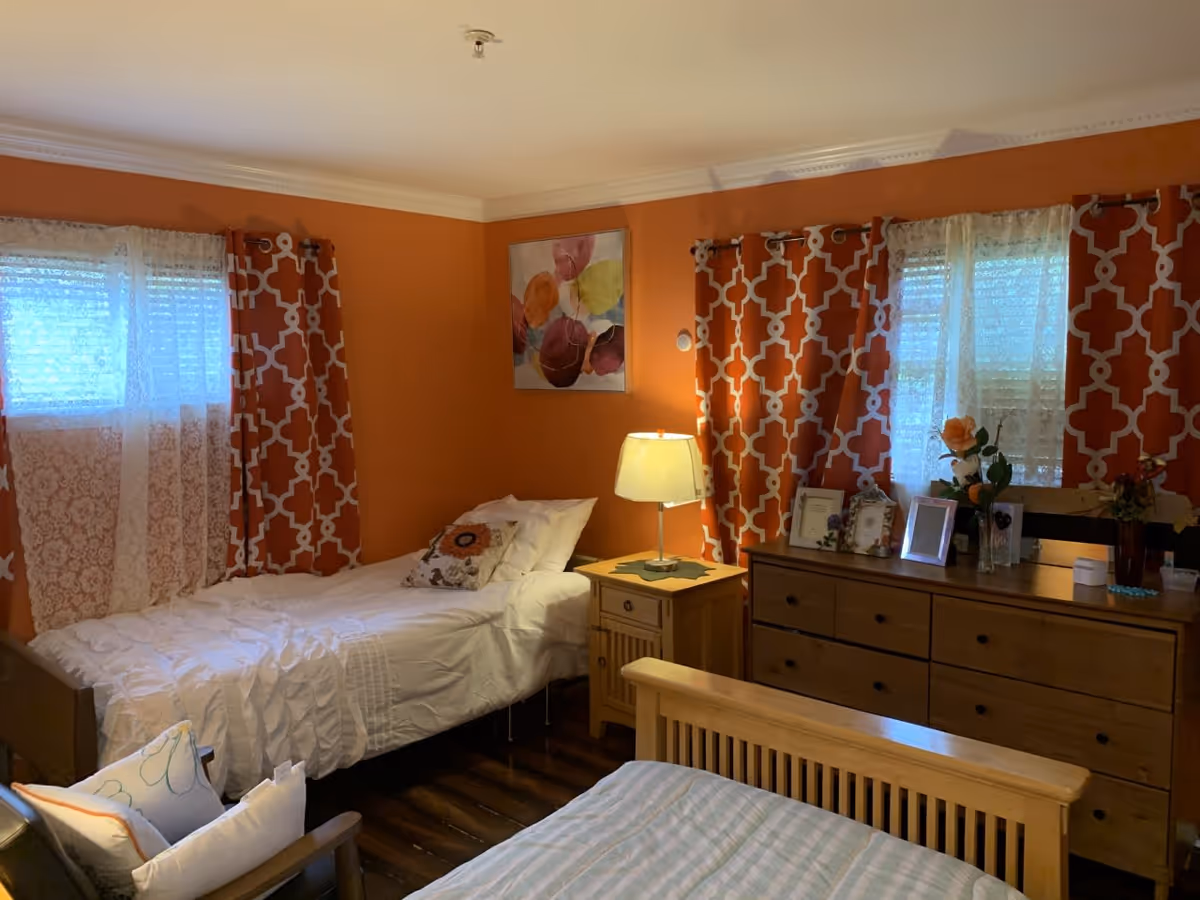 A cozy bedroom with orange walls and two windows covered with white lace curtains and orange patterned drapes. There are two beds, one with white bedding and a decorative pillow, and the other with striped bedding. A wooden nightstand with a lamp is placed between the beds. A wooden dresser with framed photos and flowers is against the wall.