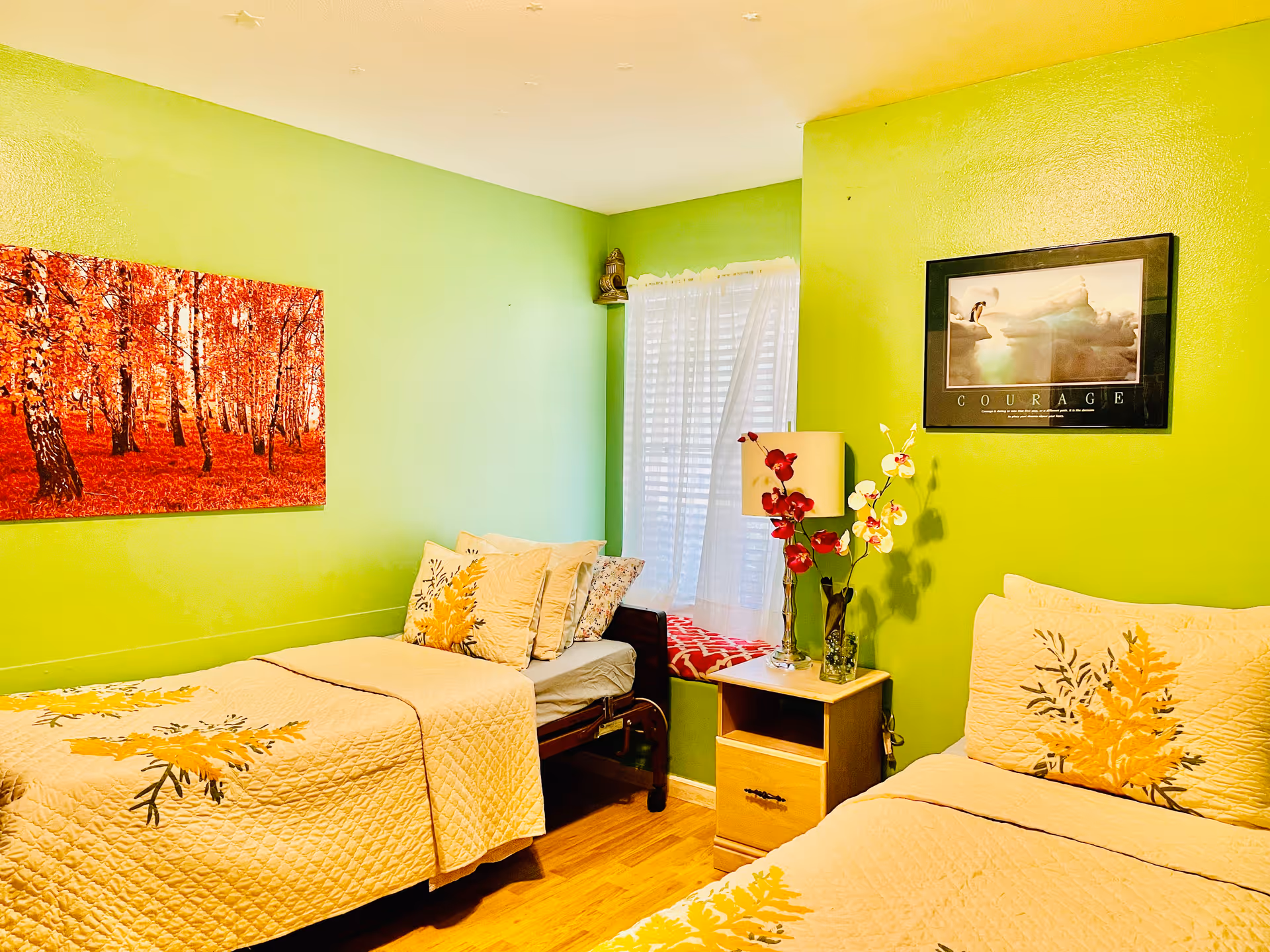 A bright bedroom with lime green walls featuring two beds with yellow floral bedspreads. Between the beds is a wooden nightstand with a lamp and a vase of red and white flowers. A window with white curtains and a red cushion is in the corner. The walls are decorated with a framed picture of a forest with red leaves and a motivational poster titled 'Courage'.