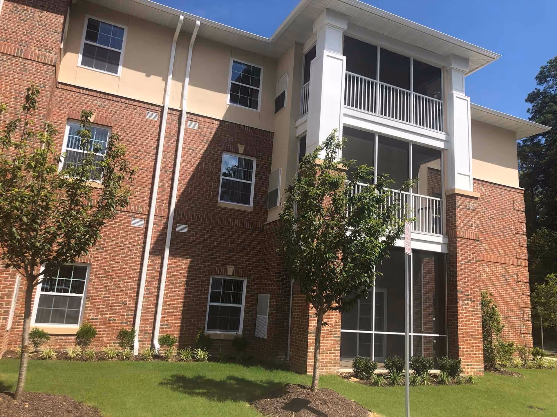 Exterior view of a three-story brick and beige building with multiple windows and screened balconies, surrounded by green grass and small trees under a clear blue sky.