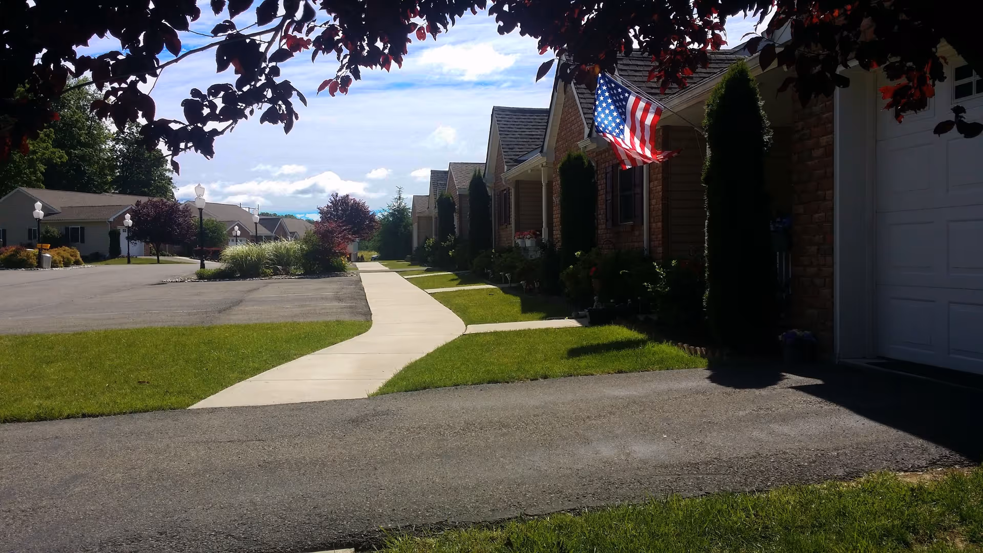 Sidewalk leading past single-story residences with an American flag, manicured lawns, and a partly cloudy sky.