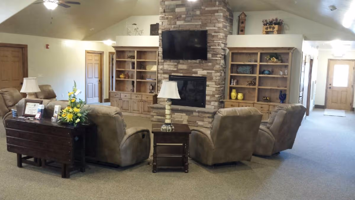 A cozy living room area in an assisted living facility featuring four beige recliner chairs arranged around a stone fireplace with a mounted flat-screen TV above it. There are two wooden bookshelves on either side of the fireplace with decorative items. A small wooden table with a lamp is placed between the chairs, and another table with a flower arrangement and framed items is behind one of the chairs. The room has beige walls, carpeted flooring, and wooden doors leading to other areas.