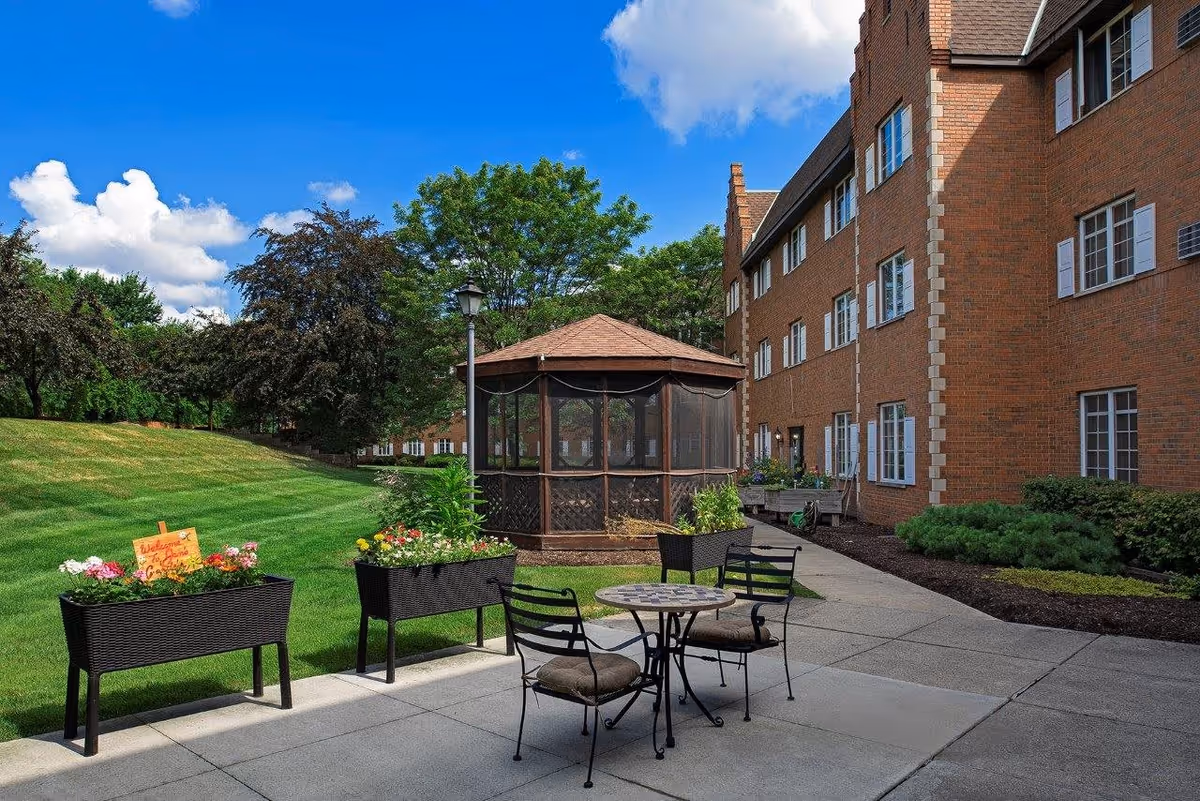 Outdoor patio area at American House East I with a small round table and four chairs on a concrete surface. There are two rectangular planters with flowers and greenery, a gazebo with a brown roof, and a brick building with multiple windows in the background. The sky is blue with some clouds, and there is a grassy area with trees and shrubs.