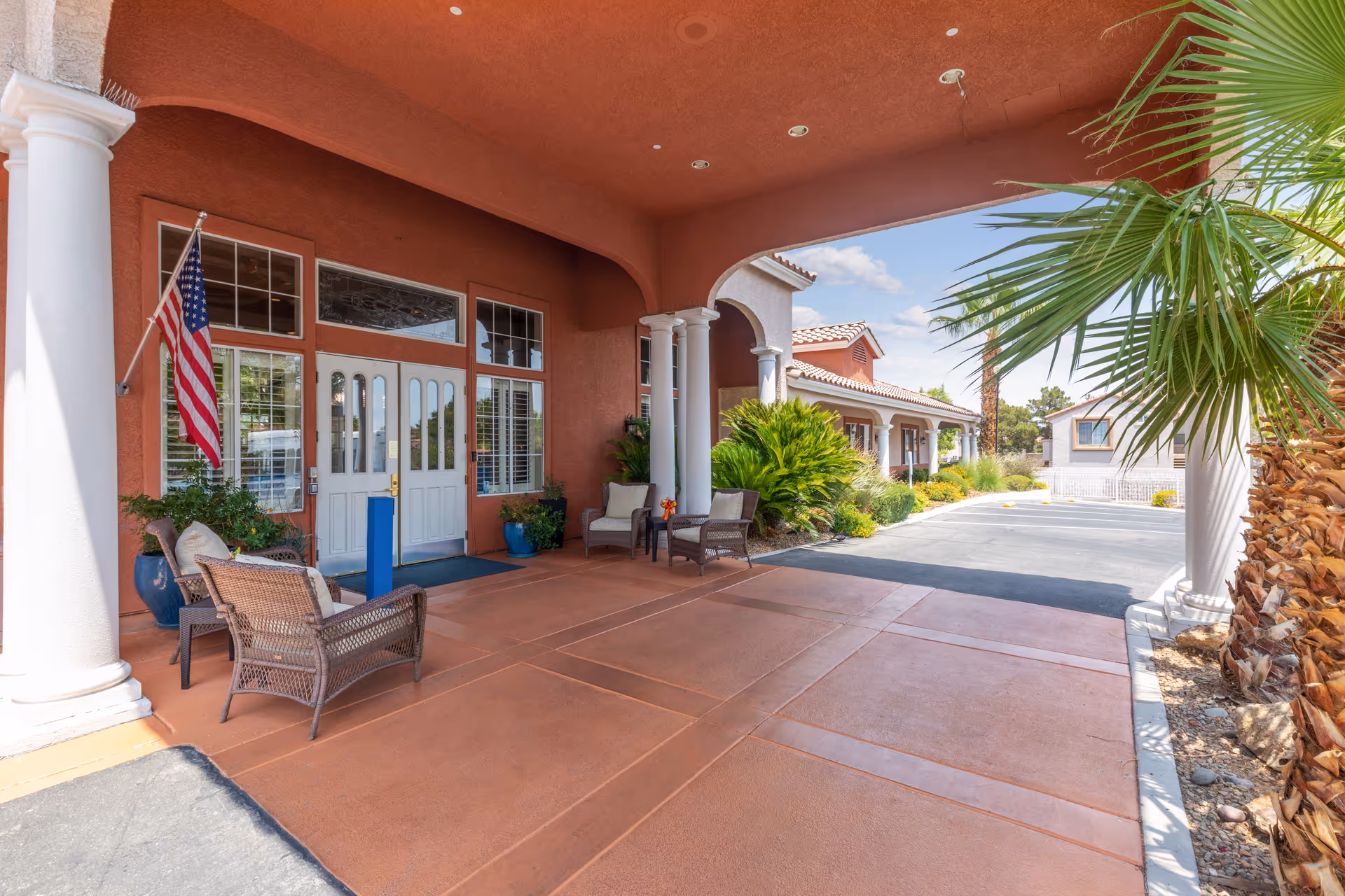 Covered entrance area of a senior living facility with white double doors, an American flag, wicker chairs with cushions, potted plants, white columns, and a driveway leading to additional buildings under a partly cloudy sky.